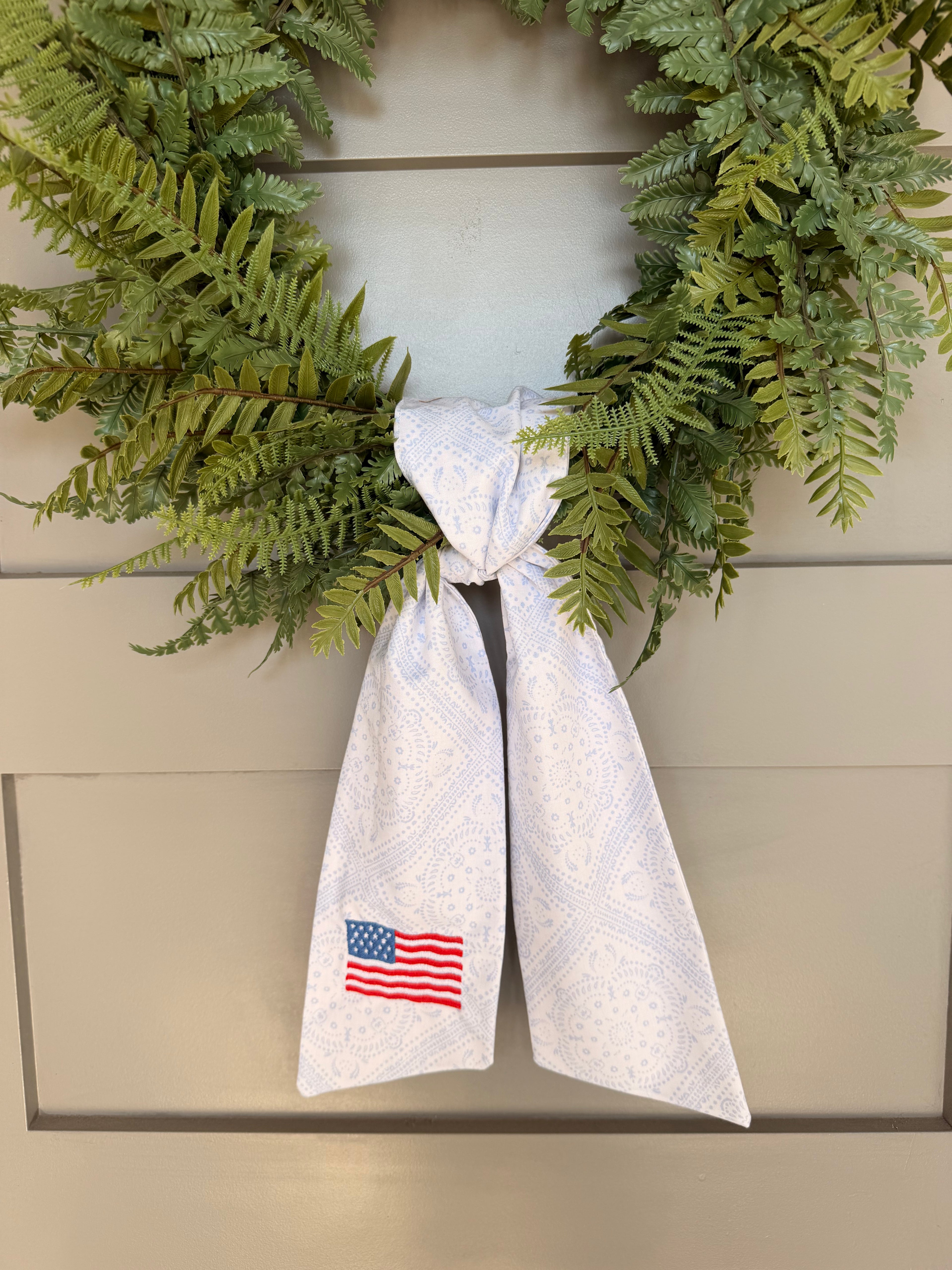A green artificial fern wreath hanging on a beige paneled door, tied at the bottom with a white paisley sash that features an American flag embroidery.