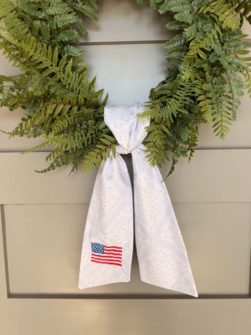 A green artificial fern wreath hanging on a beige paneled door, tied at the bottom with a white paisley sash that features an American flag embroidery.