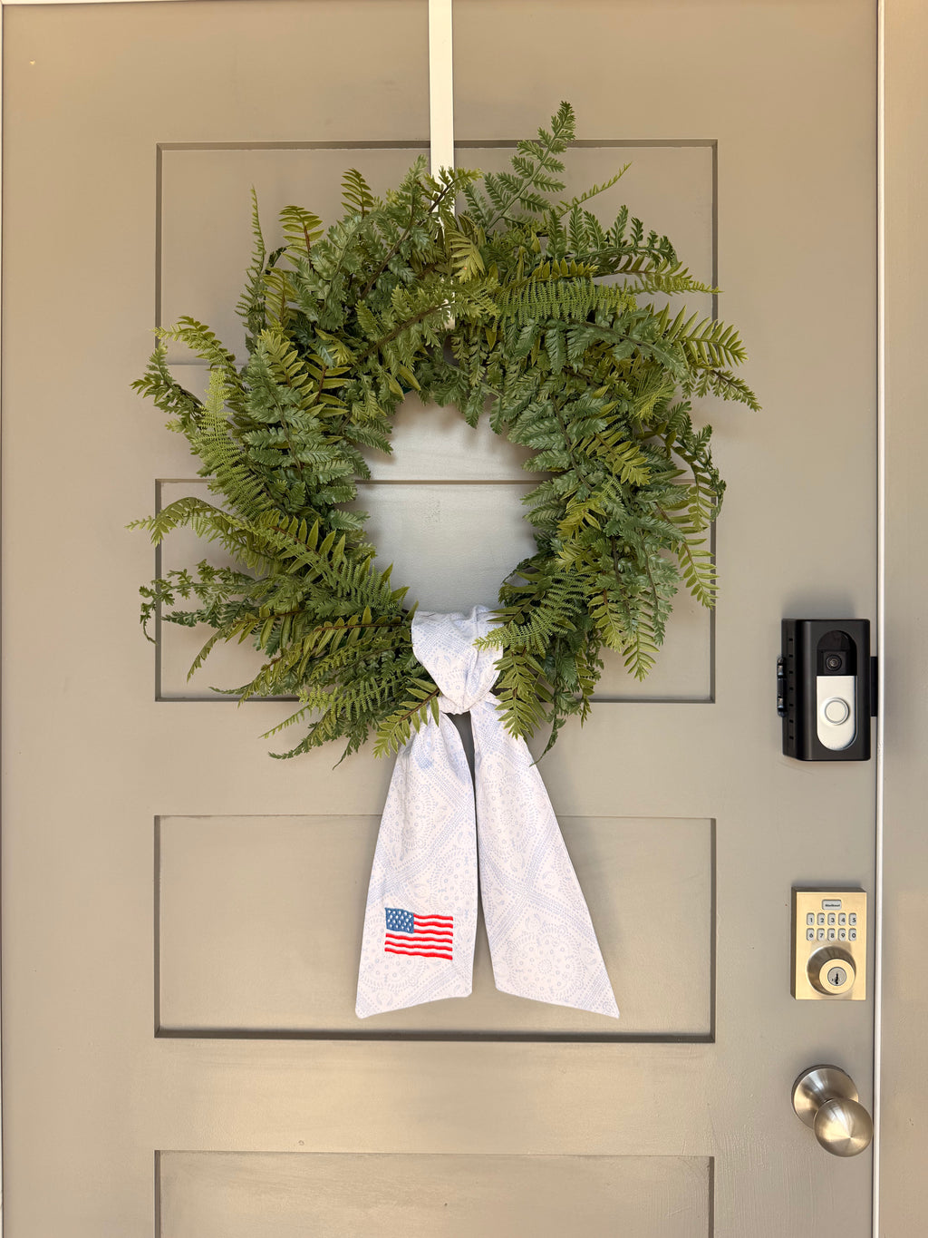 A green artificial fern wreath hanging on a beige paneled door, tied at the bottom with a white paisley sash that features an American flag embroidery.