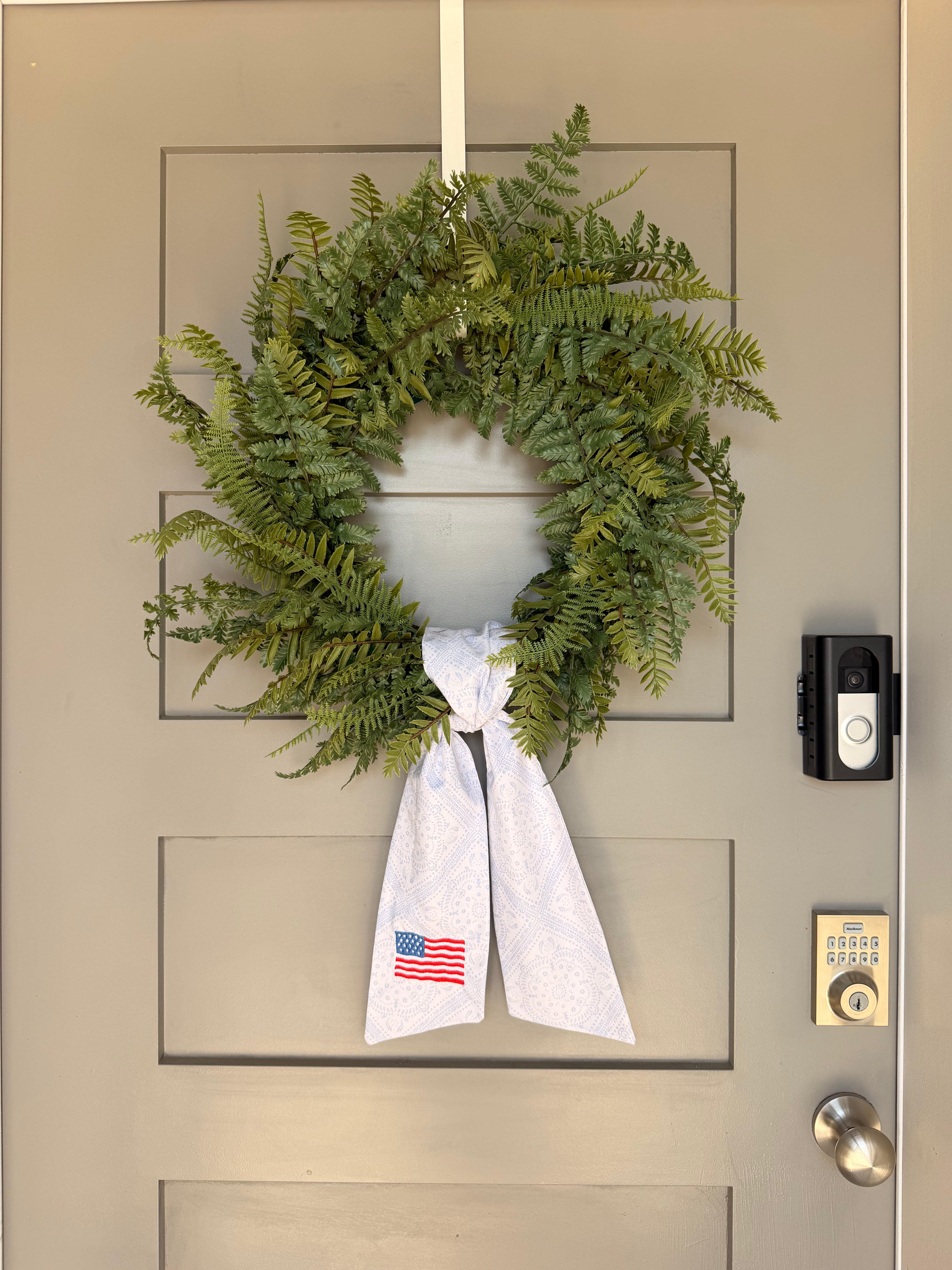 A green artificial fern wreath hanging on a beige paneled door, tied at the bottom with a white paisley sash that features an American flag embroidery.