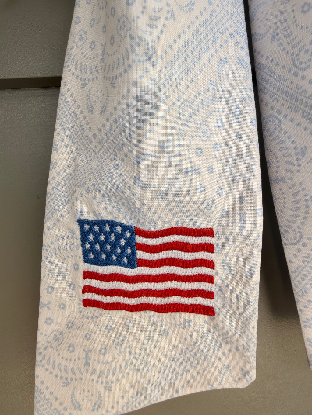 A green artificial fern wreath hanging on a beige paneled door, tied at the bottom with a white paisley sash that features an American flag embroidery.
