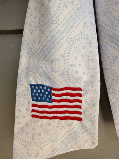 A green artificial fern wreath hanging on a beige paneled door, tied at the bottom with a white paisley sash that features an American flag embroidery.