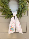 A white fabric sash with colorful confetti dots and a 'Happy Birthday' graphic embroidered, tied around a green wreath on a gray paneled door.