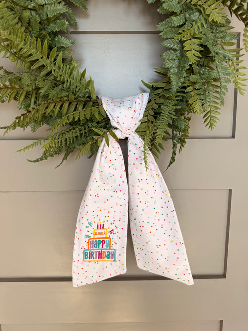A white fabric sash with colorful confetti dots and a 'Happy Birthday' graphic embroidered, tied around a green wreath on a gray paneled door.