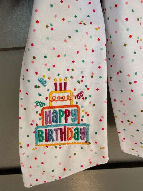 A white fabric sash with colorful confetti dots and a 'Happy Birthday' graphic embroidered, tied around a green wreath on a gray paneled door.