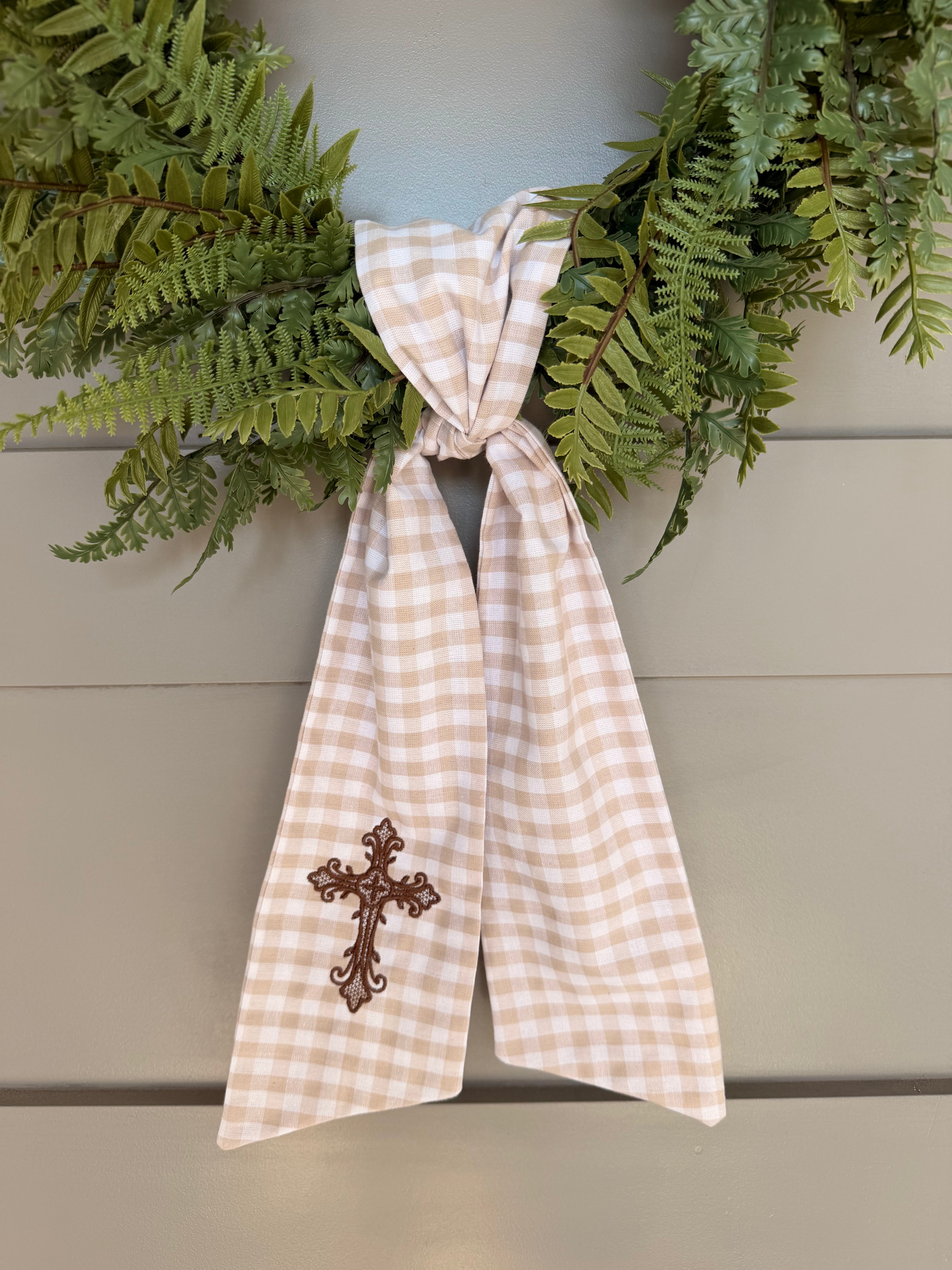 A beige and white gingham wreath sash with a brown embroidered cross, tied around a green fern wreath on a gray paneled door.