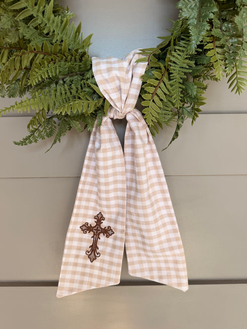 A beige and white gingham wreath sash with a brown embroidered cross, tied around a green fern wreath on a gray paneled door.