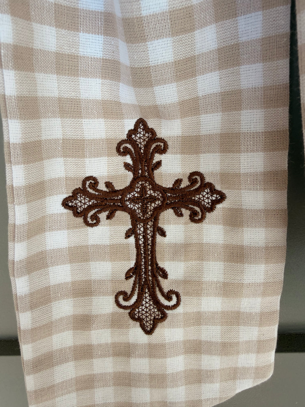A beige and white gingham wreath sash with a brown embroidered cross, tied around a green fern wreath on a gray paneled door.