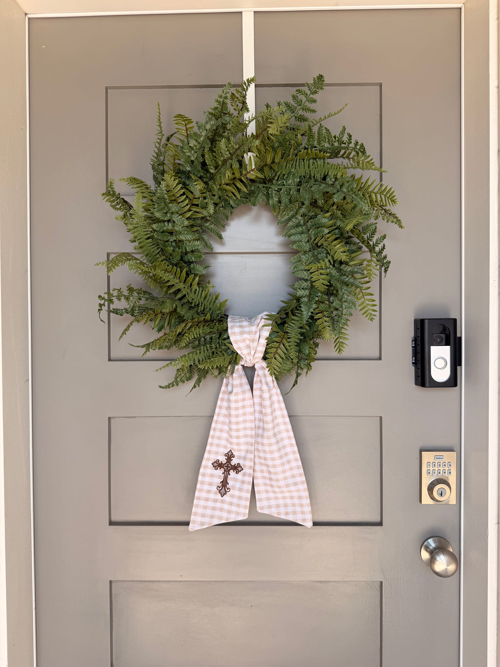 A beige and white gingham wreath sash with a brown embroidered cross, tied around a green fern wreath on a gray paneled door.