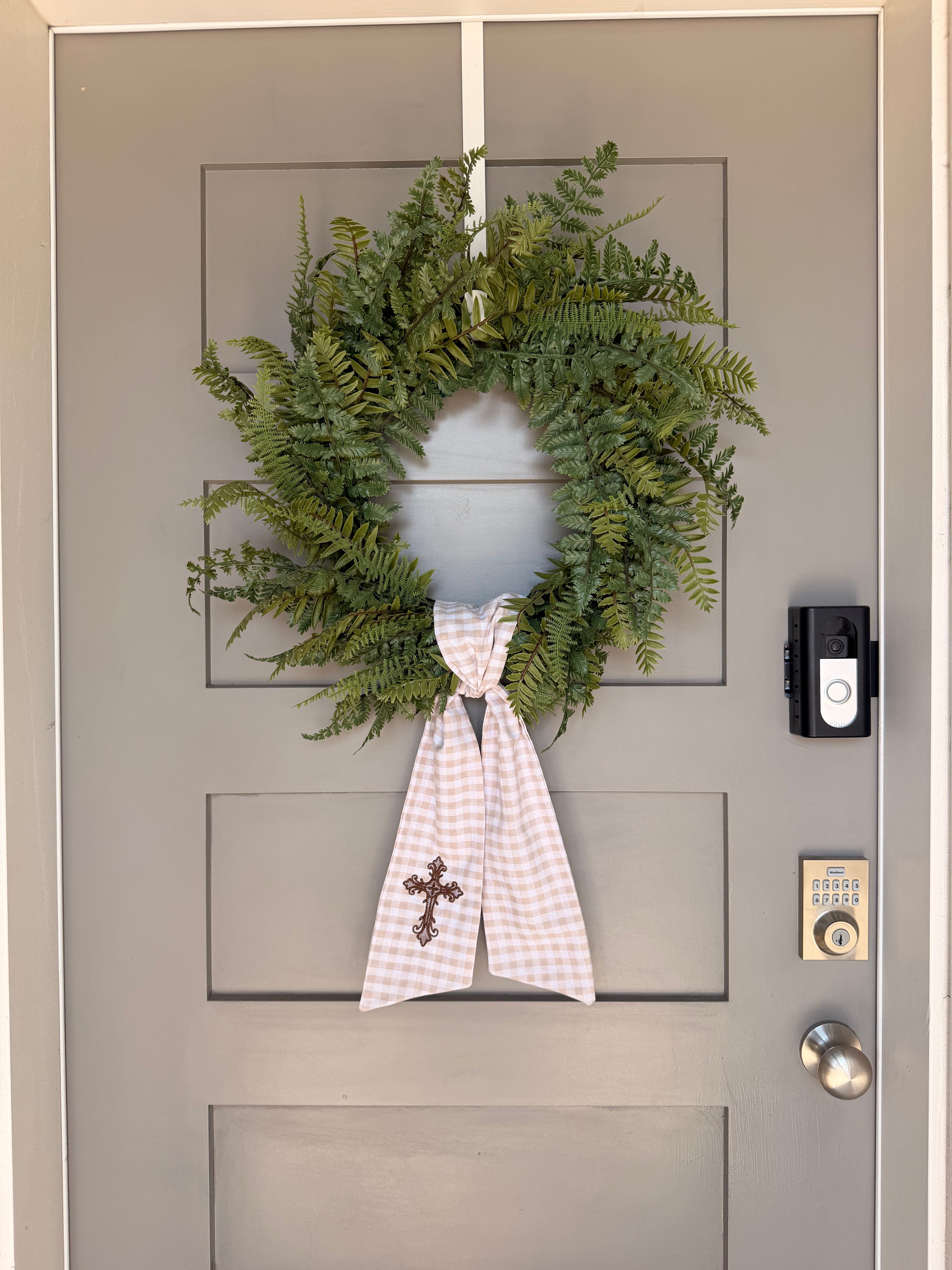 A beige and white gingham wreath sash with a brown embroidered cross, tied around a green fern wreath on a gray paneled door.
