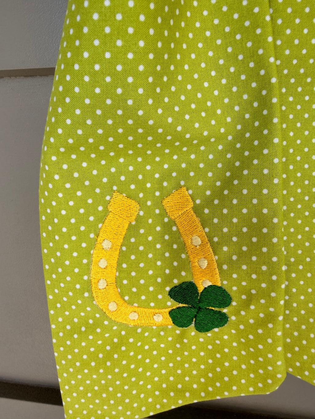 A green fabric sash with white polka dots and St. Patrick's Day themed embroidery (a horseshoe and a four-leaf clover) hanging from a green faux fern wreath on a light gray paneled door.