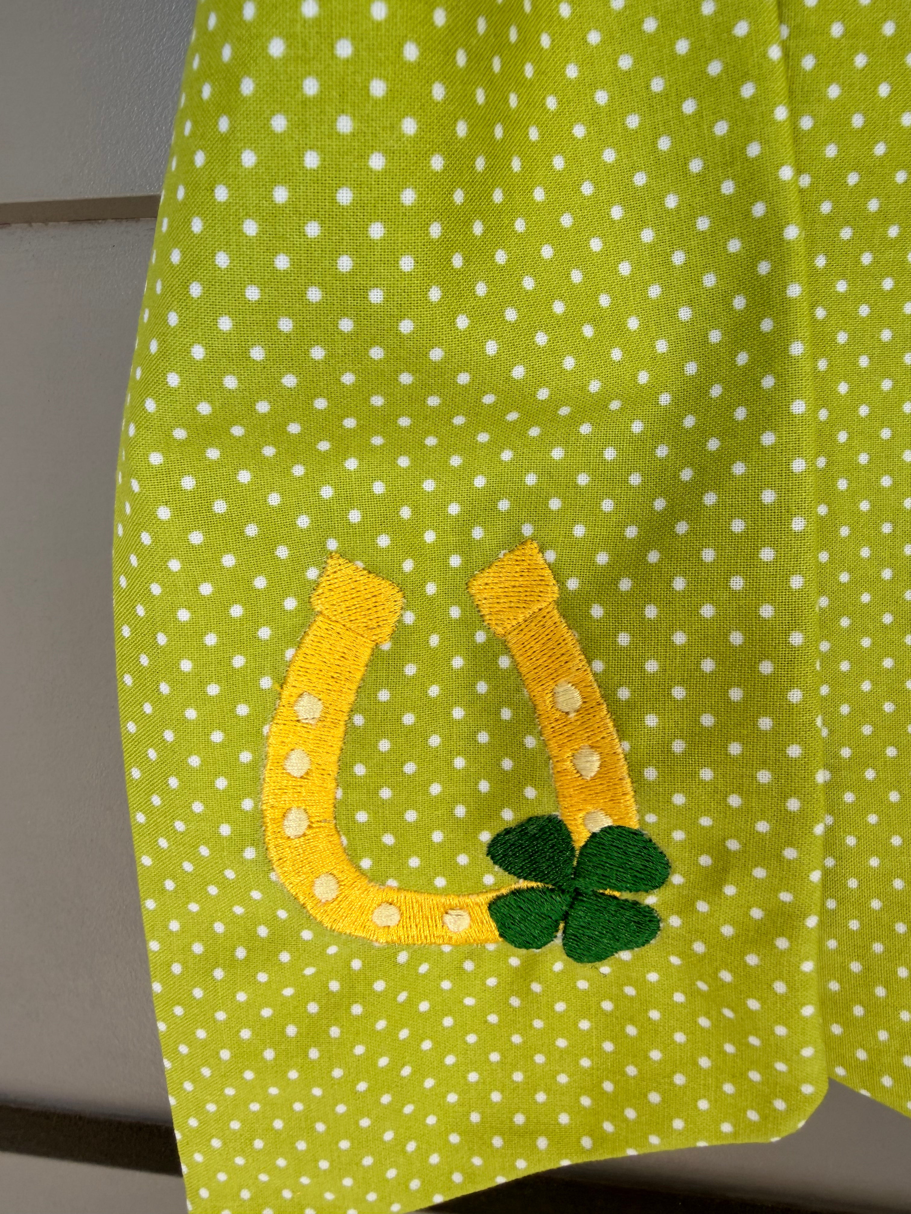 A green fabric sash with white polka dots and St. Patrick's Day themed embroidery (a horseshoe and a four-leaf clover) hanging from a green faux fern wreath on a light gray paneled door.