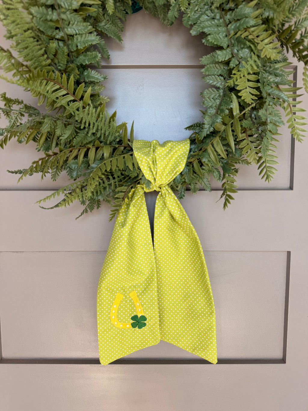 A green fabric sash with white polka dots and St. Patrick's Day themed embroidery (a horseshoe and a four-leaf clover) hanging from a green faux fern wreath on a light gray paneled door.