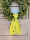 A green fabric sash with white polka dots and St. Patrick's Day themed embroidery (a horseshoe and a four-leaf clover) hanging from a green faux fern wreath on a light gray paneled door.