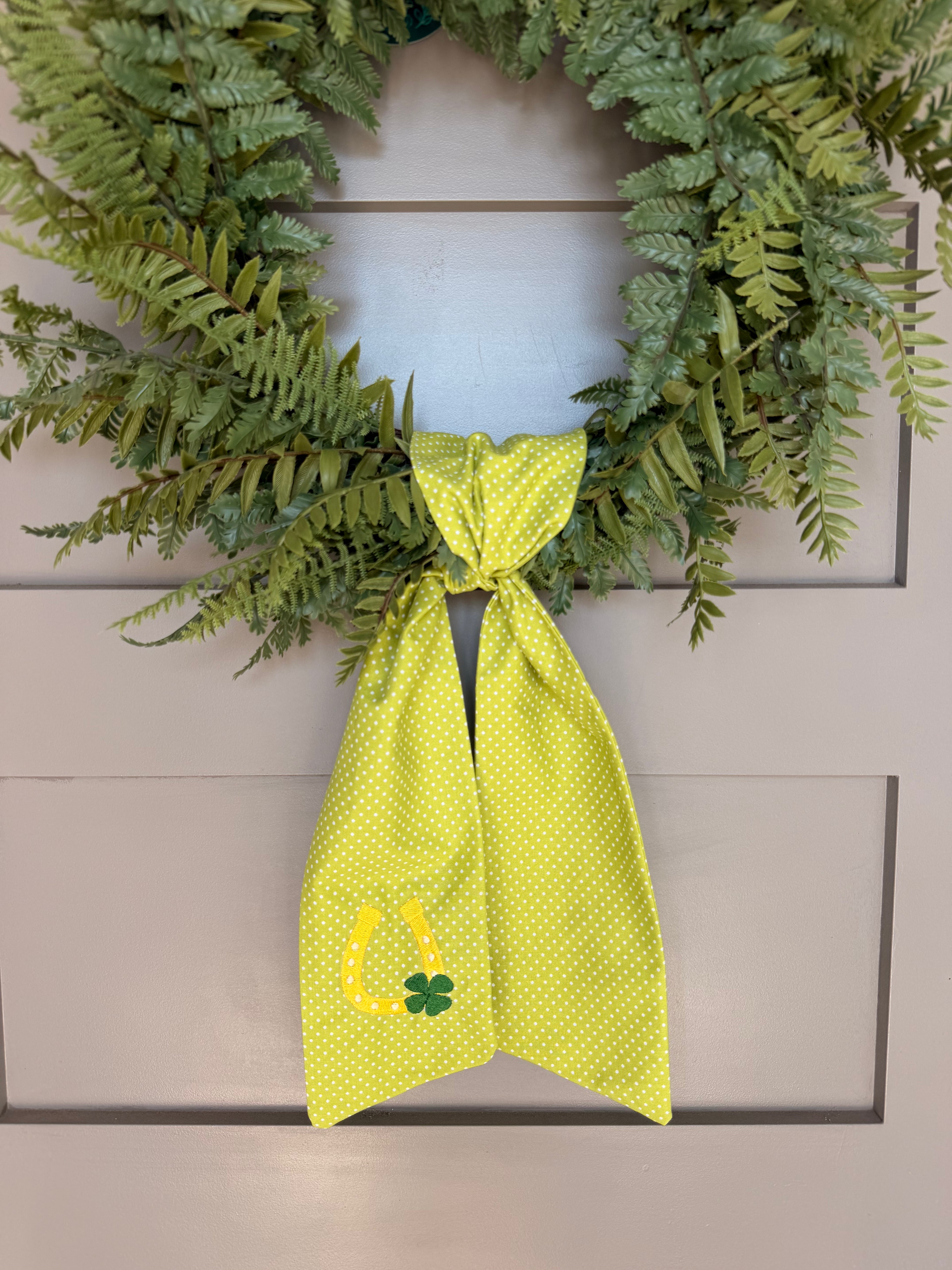 A green fabric sash with white polka dots and St. Patrick's Day themed embroidery (a horseshoe and a four-leaf clover) hanging from a green faux fern wreath on a light gray paneled door.