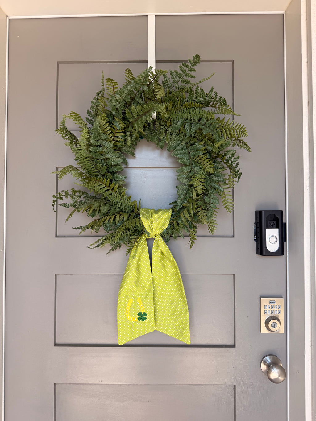 A green fabric sash with white polka dots and St. Patrick's Day themed embroidery (a horseshoe and a four-leaf clover) hanging from a green faux fern wreath on a light gray paneled door.