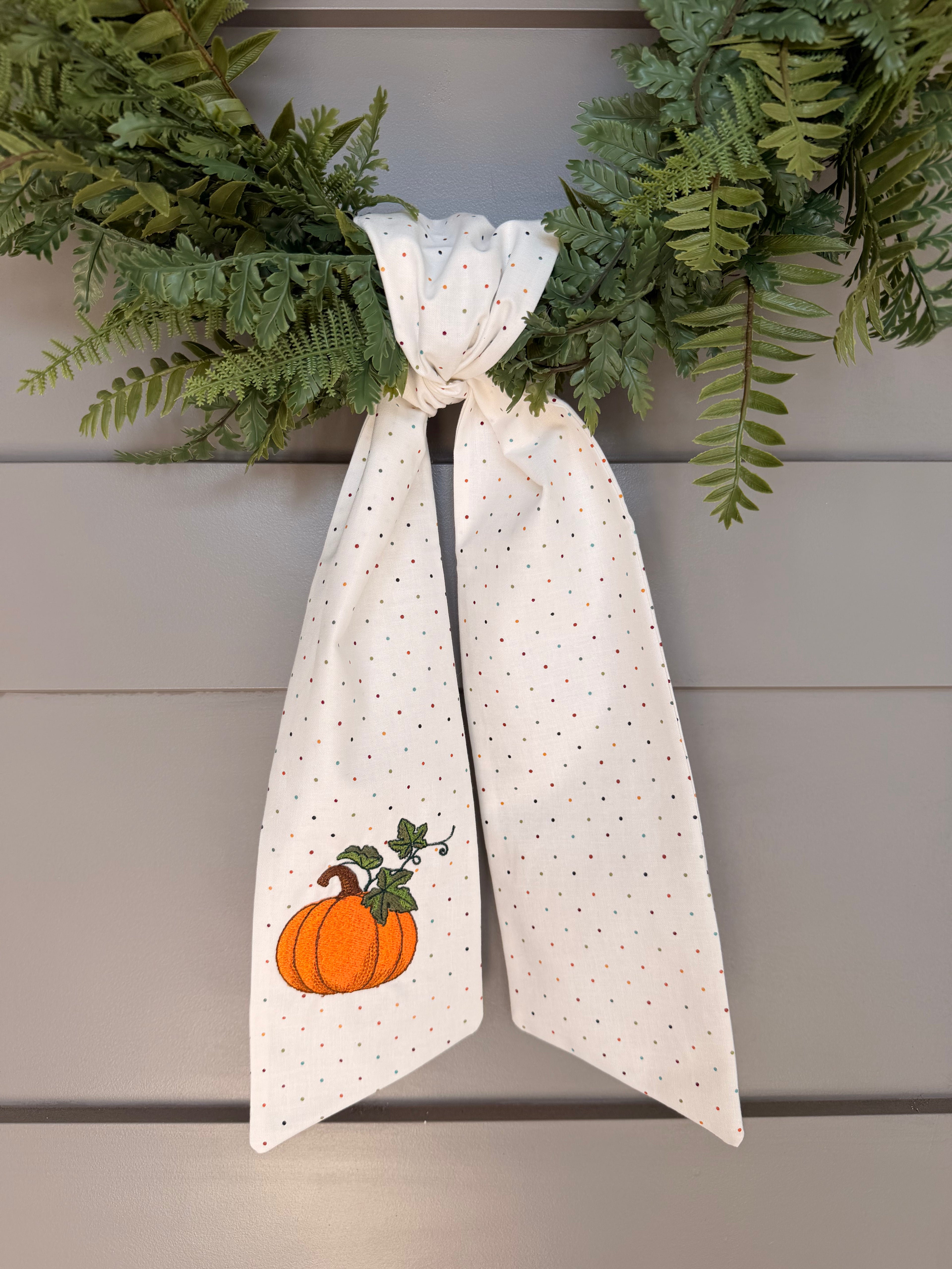 A white fabric sash with tiny, multi-color polka dots and an orange pumpkin embroidered, tied in a knot and hung on a green wreath.