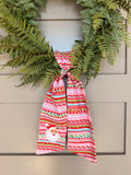 A festive wreath made of green ferns, decorated with a large red, white, and green striped bow featuring a small embroidered Santa Claus face, hanging on a grey paneled door.