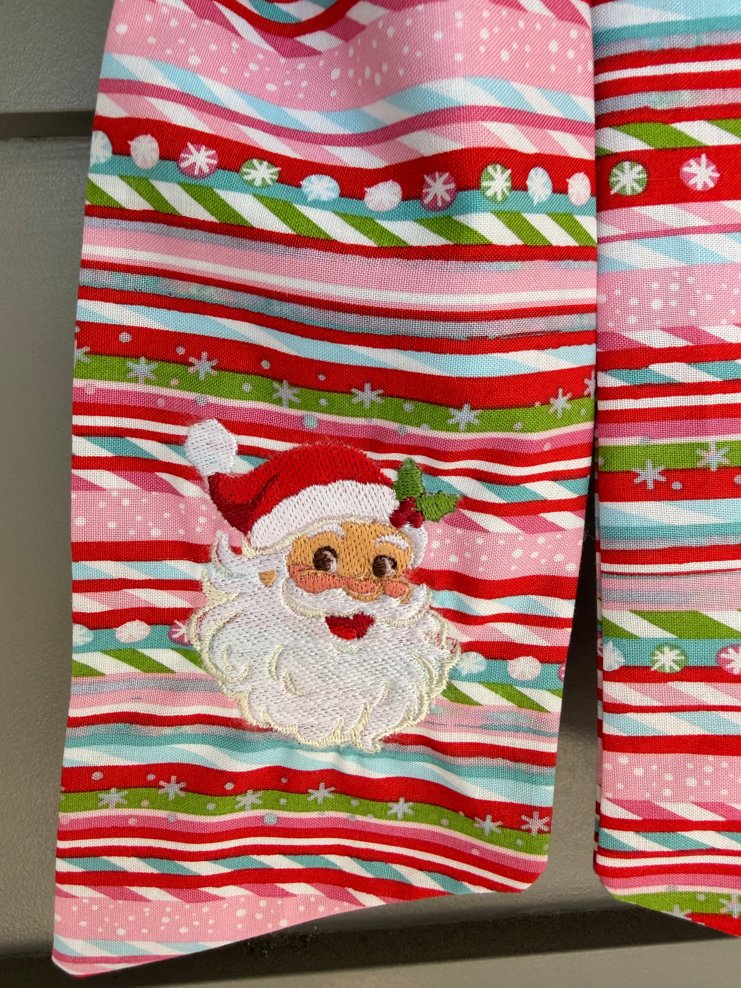 A festive wreath made of green ferns, decorated with a large red, white, and green striped bow featuring a small embroidered Santa Claus face, hanging on a grey paneled door.