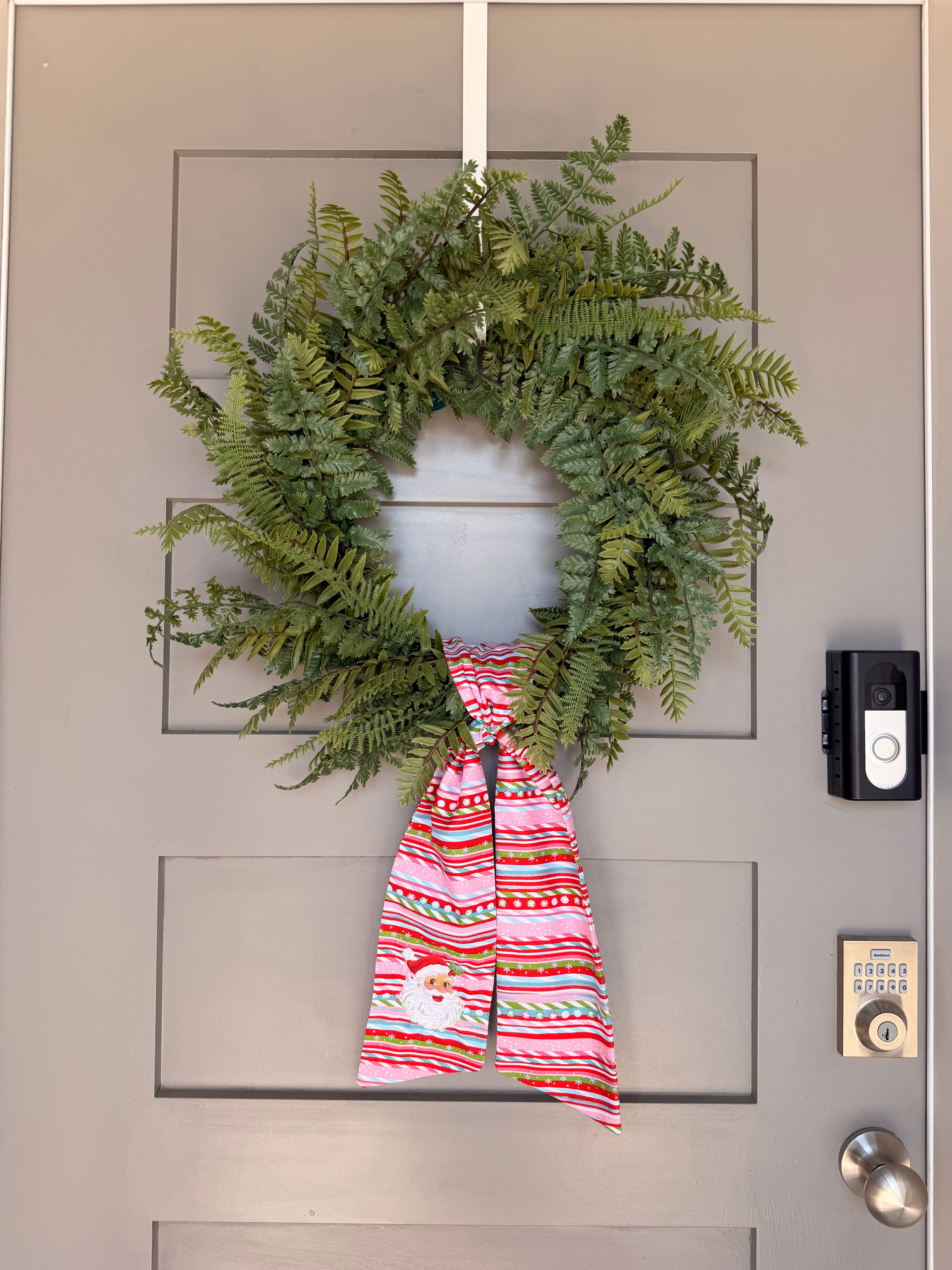 A festive wreath made of green ferns, decorated with a large red, white, and green striped bow featuring a small embroidered Santa Claus face, hanging on a grey paneled door.