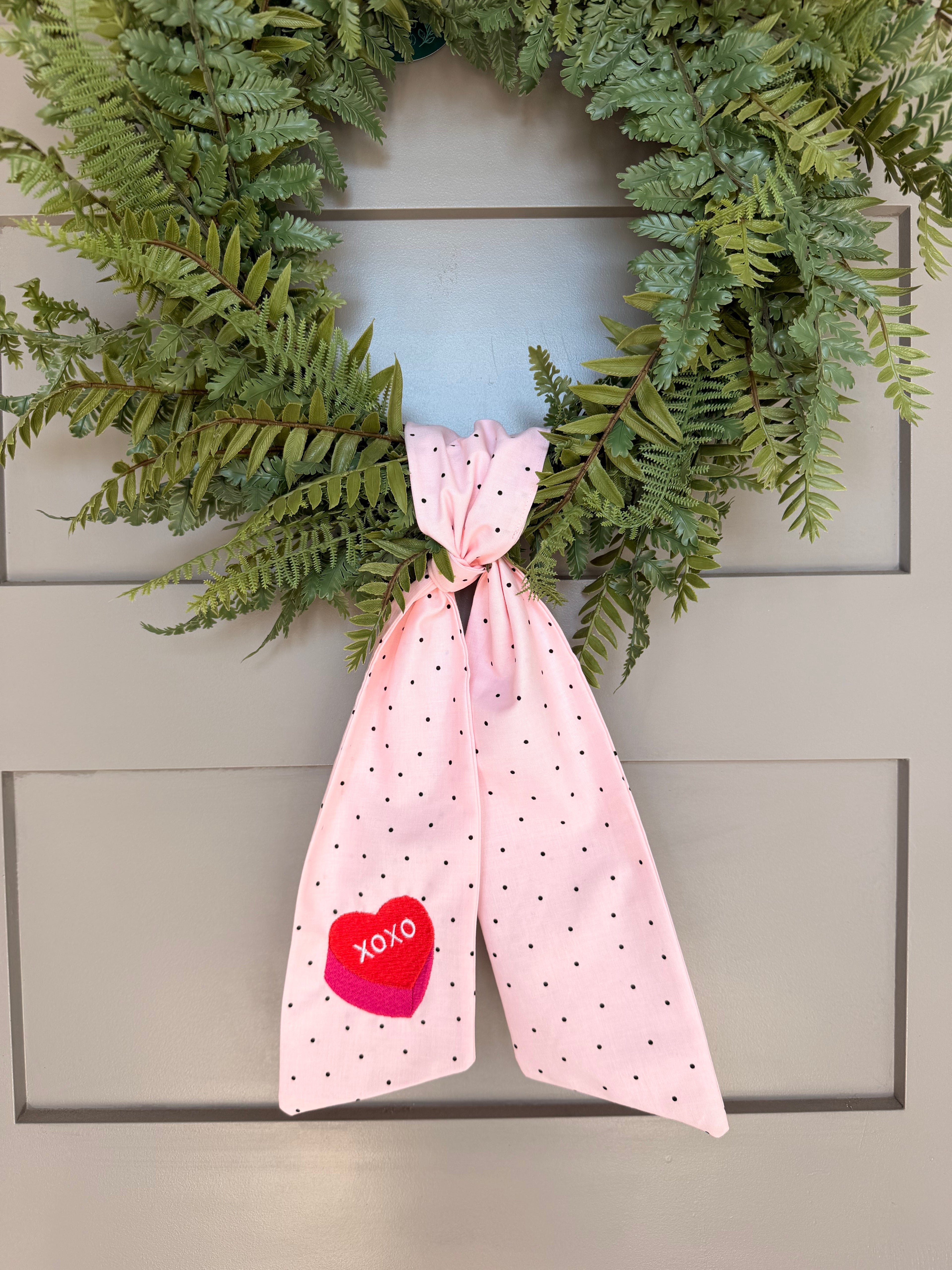 A pink fabric Valentine's Day wreath sash with small black polka dots and a red heart patch that says 'XOXO', hanging from a green wreath on a gray paneled door.