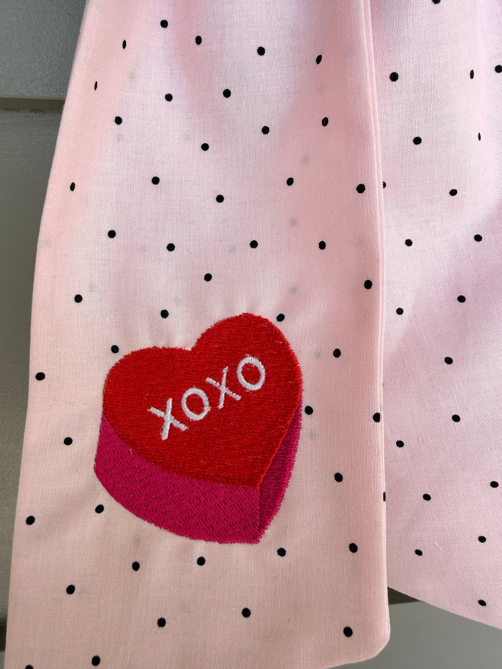 A pink fabric Valentine's Day wreath sash with small black polka dots and a red heart patch that says 'XOXO', hanging from a green wreath on a gray paneled door.
