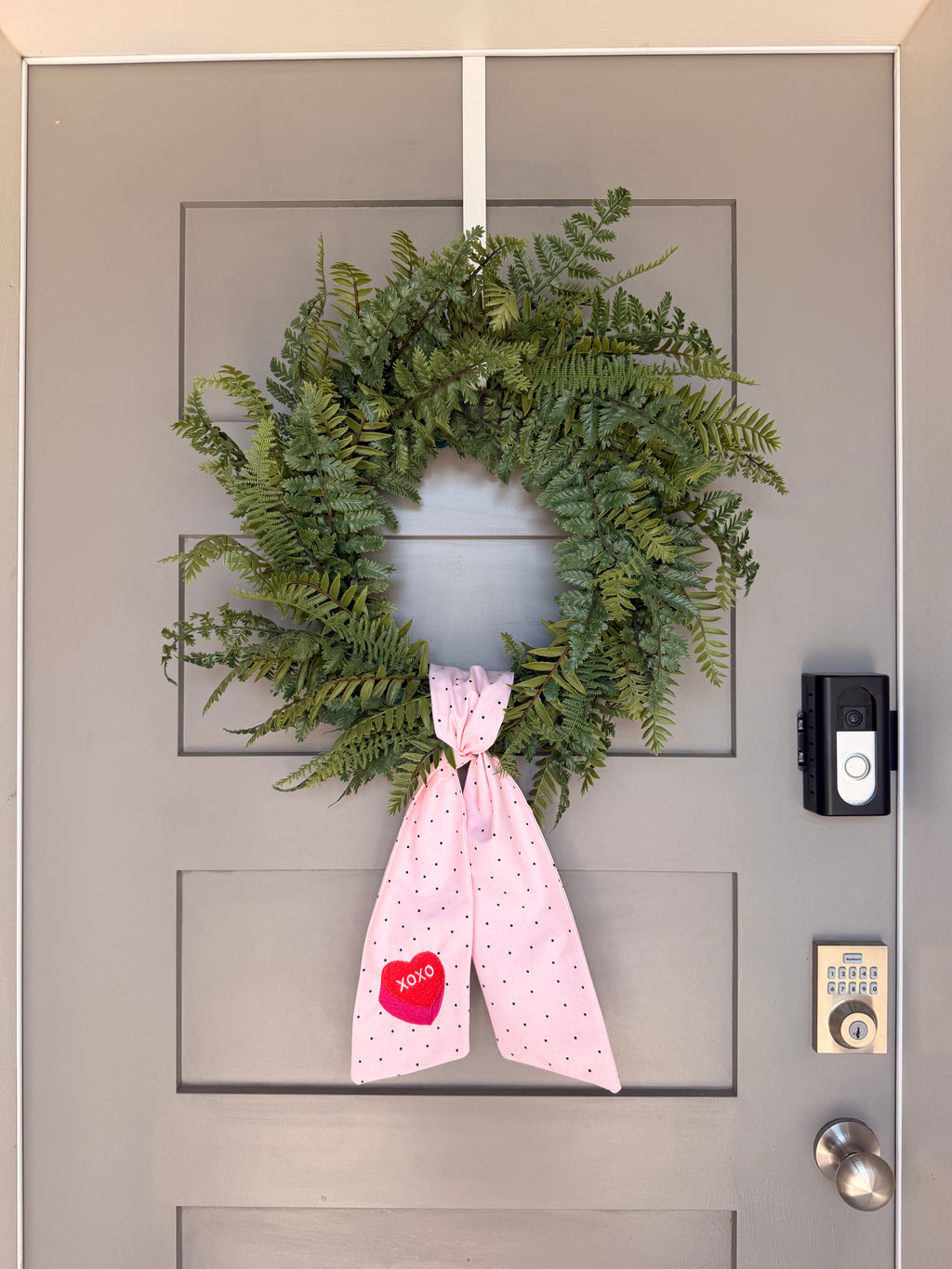 A pink fabric Valentine's Day wreath sash with small black polka dots and a red heart patch that says 'XOXO', hanging from a green wreath on a gray paneled door.